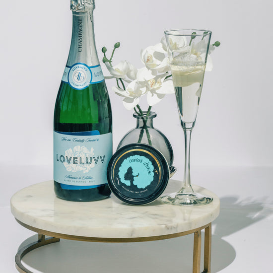 Bottle of LOVELUVV champagne, a one ounce tin of Classic Osetra caviar and a champagne glass with white orchids on a marble tray.