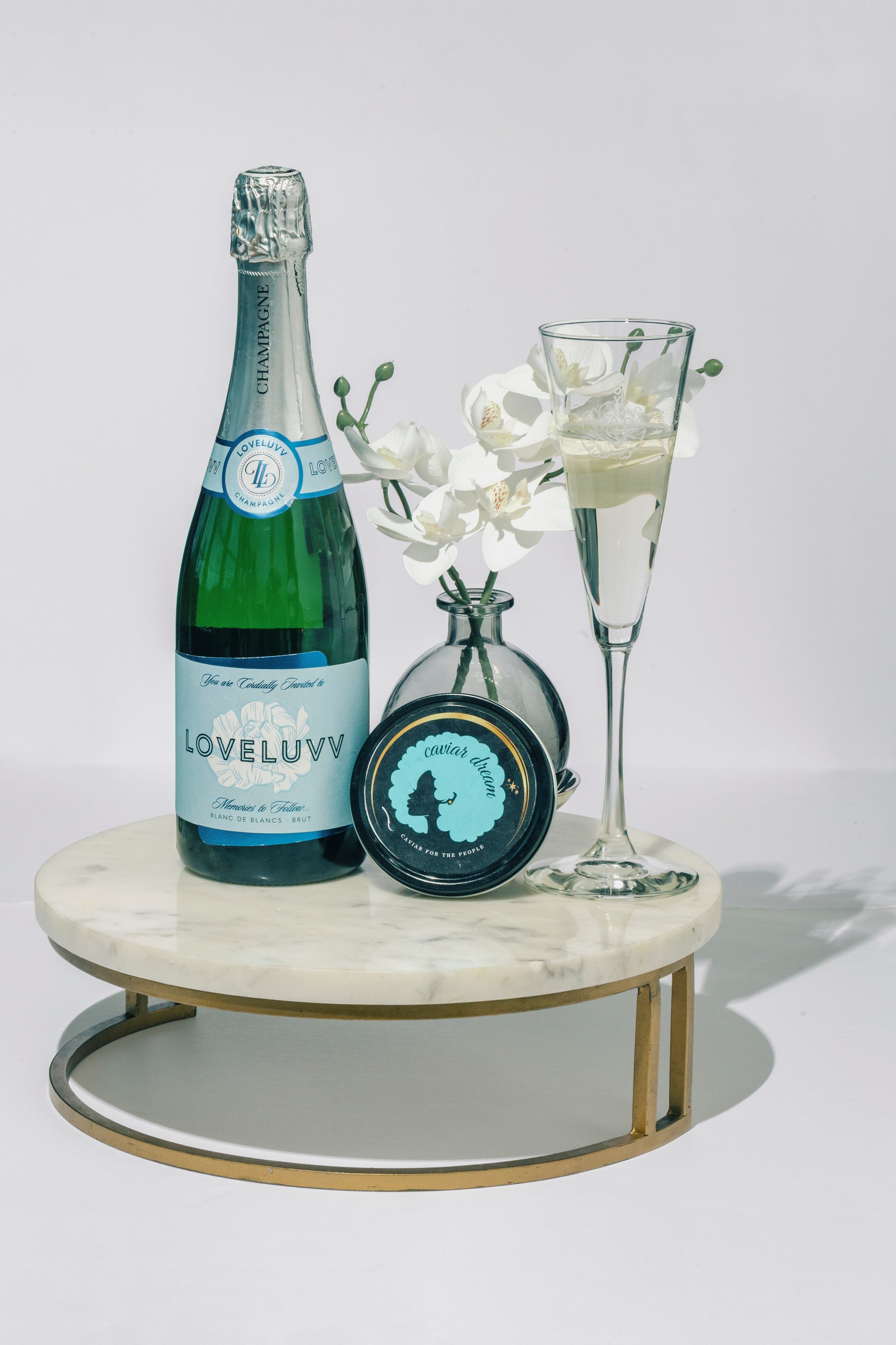 Bottle of LOVELUVV champagne, a one ounce tin of Classic Osetra caviar and a champagne glass with white orchids on a marble tray.