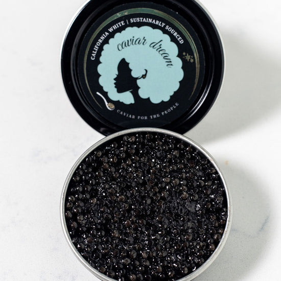 An open tin of dark brown, almost black California White caviar with a visible Caviar Dream logo on the top of the tin. Both on a white background