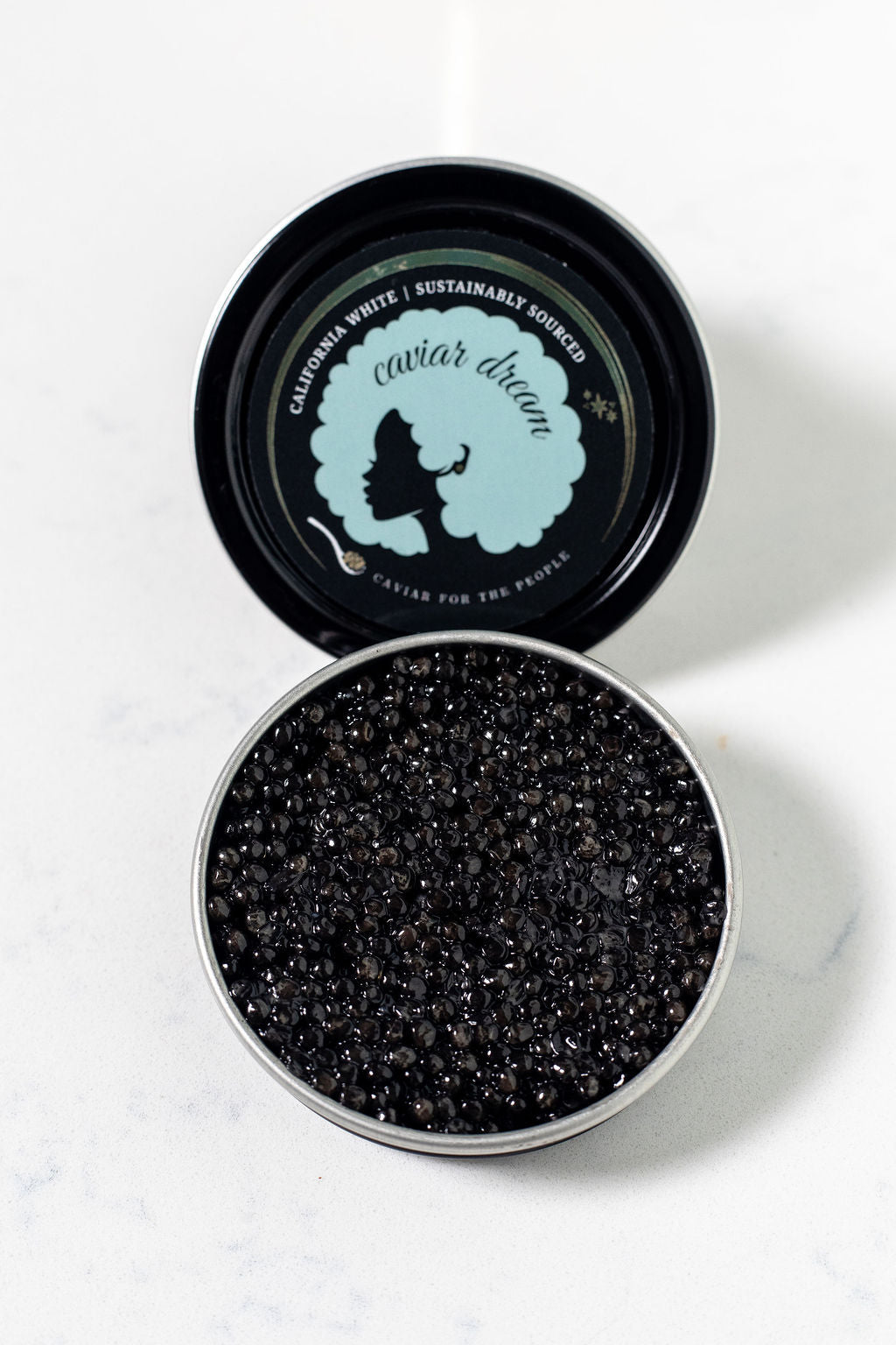 An open tin of dark brown, almost black California White caviar with a visible Caviar Dream logo on the top of the tin. Both on a white background