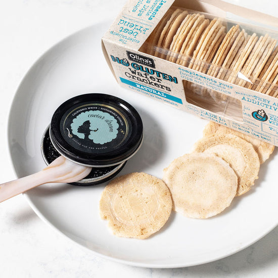 Box of Olina's gluten-free crackers, more crackers on a white plate with a white background, alongside an open tin of California White caviar and a mother of pearl spoon.