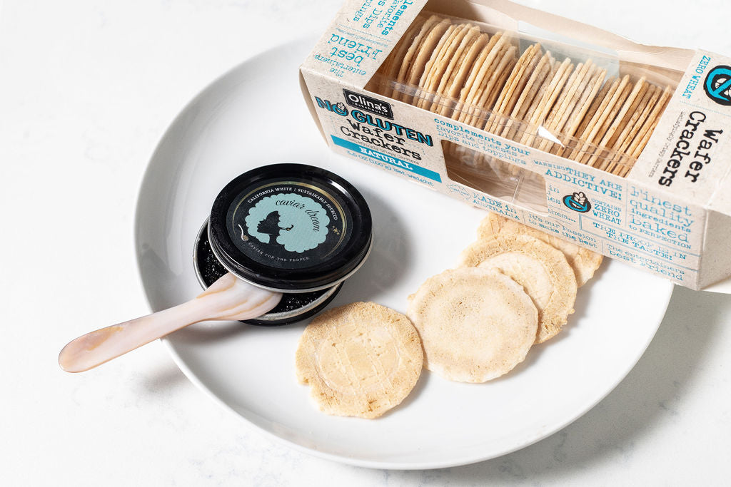 Box of Olina's gluten-free crackers, more crackers on a white plate with a white background, alongside an open tin of California White caviar and a mother of pearl spoon.