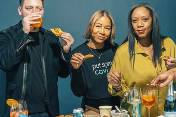 Three people standing together at a table, each holding a piece of pizza and drinking from a glass, with food and drinks on the table against a dark blue background. The person in the center wears a black sweatshirt with the text 'put some cavi' in white and light blue.
