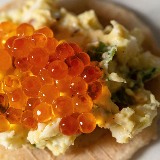 Close-up of an Olina's gluten-free cracker topped with egg salad and smoked trout roe on a white background.