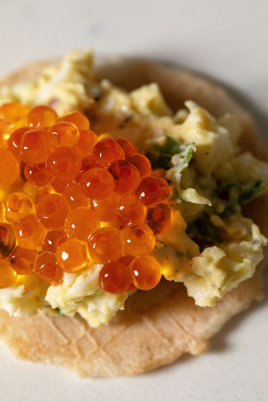 Close-up of an Olina's gluten-free cracker topped with egg salad and smoked trout roe on a white background.