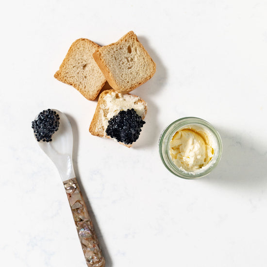 Three crunchy, mini toasts with caviar, a spoon with caviar, and a jar of creme fraiche on a white background