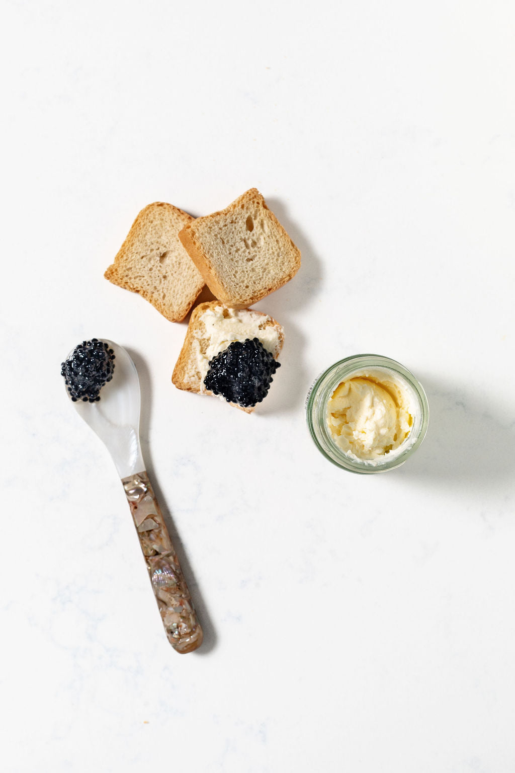 Three crunchy, mini toasts with caviar, a spoon with caviar, and a jar of creme fraiche on a white background